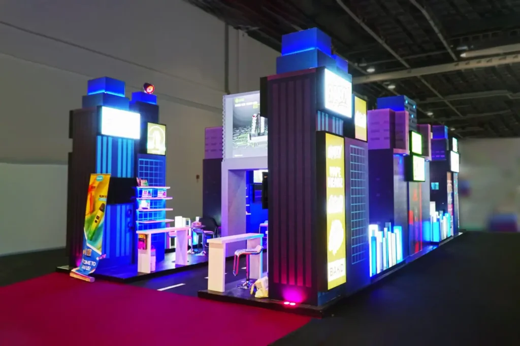 Illuminated cityscape exhibition stand featuring tall display towers, LED branding panels, and interactive product showcase