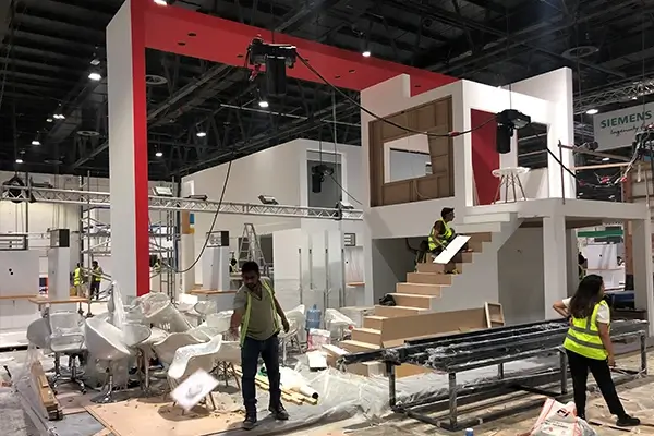 Exhibition booth fit-out in progress with workers installing structural elements, staircase, and interior components